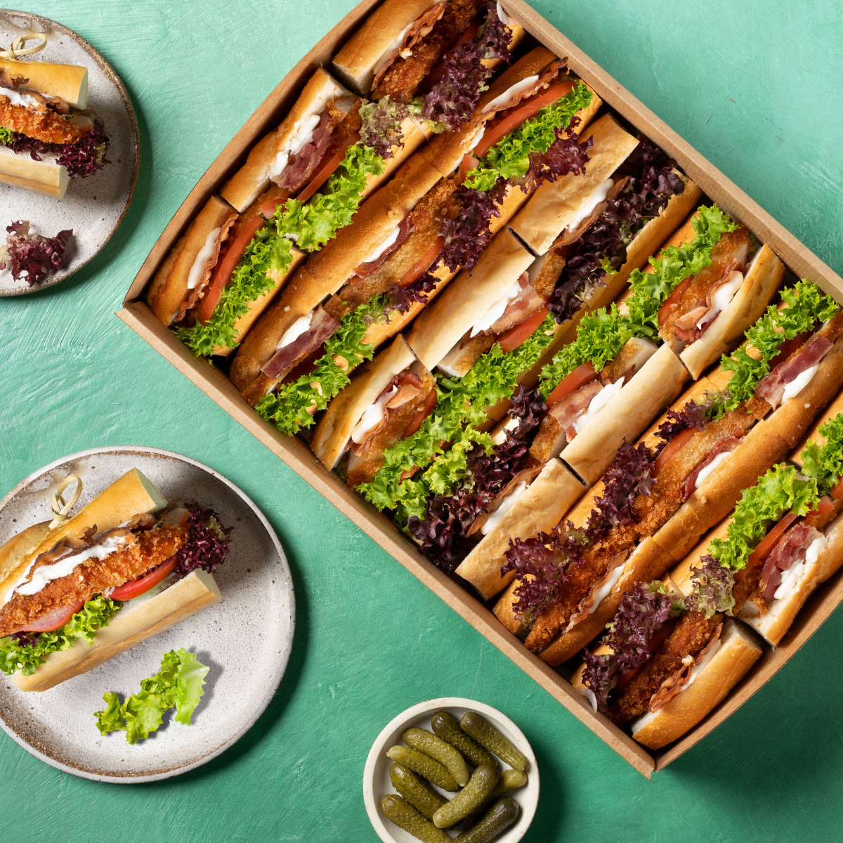 Box of sub sandwiches with plates and pickles on a green background