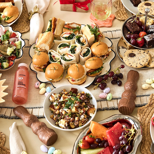 Diverse food platter with sliders, salads, fruits, and a bottle of wine on a patterned tablecloth.
